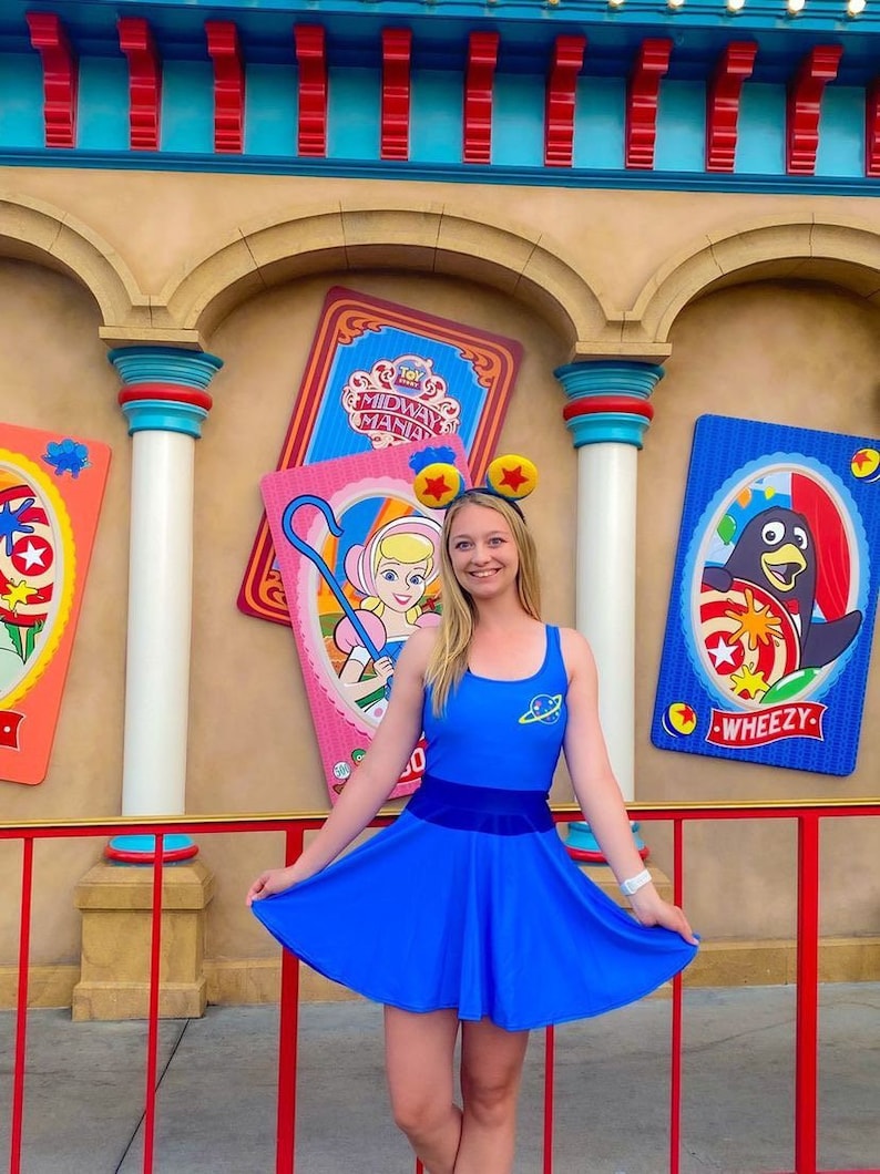 May include: A woman wearing a blue dress with a space-themed design and a pair of red Mickey Mouse ears stands in front of a colorful wall with Toy Story Midway Mania signs.