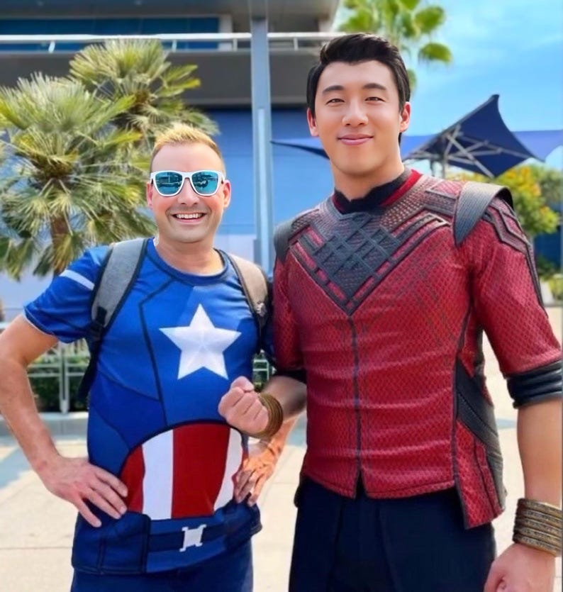 May include: Two people are standing next to each other. The person on the left is wearing a blue, white, and red t-shirt with a star and a circle design. The person on the right is wearing a red and black costume with a geometric pattern.