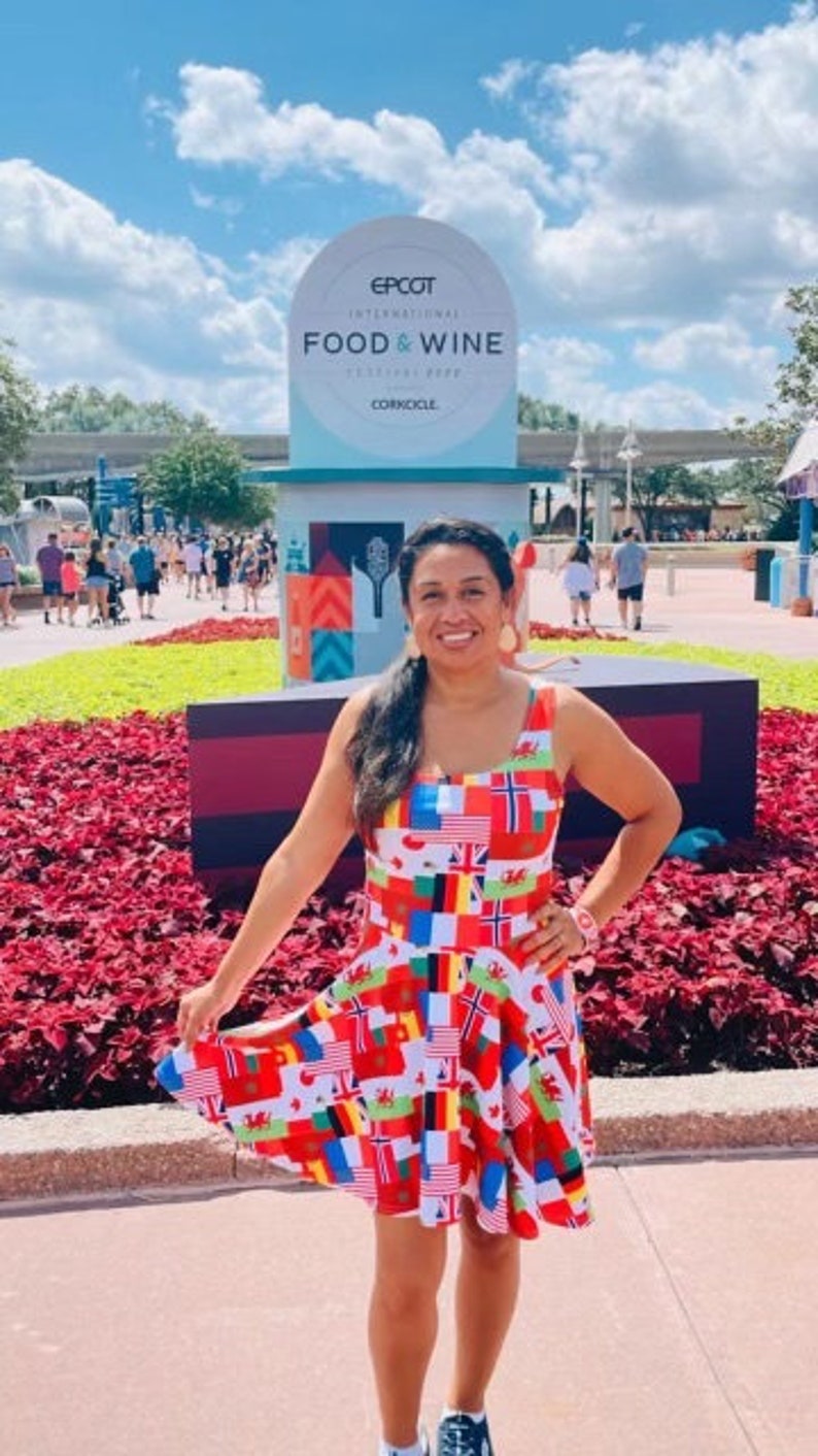 Puede incluir: Un vestido sin mangas con un estampado integral de varias banderas internacionales en colores vibrantes que incluyen rojo, blanco, azul, amarillo y verde. El vestido tiene un escote redondo y una falda acampanada que llega por encima de la rodilla.