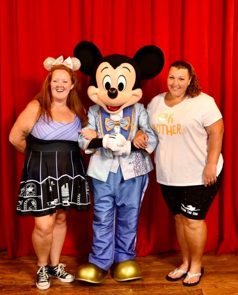 May include: Mickey Mouse, dressed in a blue suit with a gold bow tie, poses with two people. The person on the left is wearing a purple and black dress with a white bow in her hair. The person on the right is wearing a white t-shirt with the text "Oh Brother" and black shorts with the text "Under The Sea".