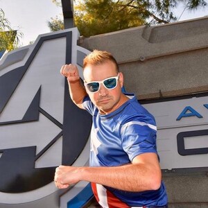 May include: A man wearing a blue and red superhero shirt with a white star design, poses in front of a large white and gray "A" logo. He is wearing blue-tinted sunglasses and has a determined expression on his face.