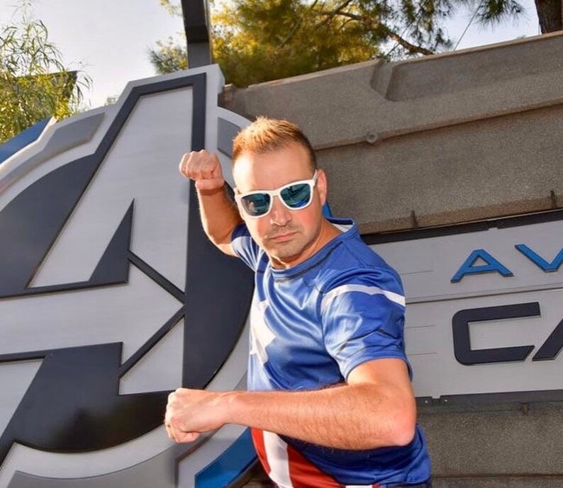 May include: A man wearing a blue and red superhero shirt with a white star design, poses in front of a large white and gray "A" logo. He is wearing blue-tinted sunglasses and has a determined expression on his face.