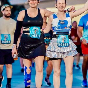 May include: Two women wearing running gear and race numbers. The woman on the left is wearing a black sports bra, black shorts, and a blue knee brace. The woman on the right is wearing a white and blue R2-D2 themed tank top, a silver sequined skirt, and a blue race number. Both women are smiling and appear to be enjoying themselves.