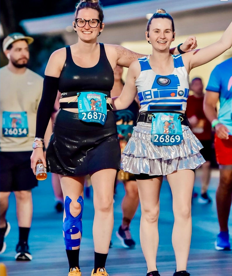 May include: Two women wearing running gear and race numbers. The woman on the left is wearing a black sports bra, black shorts, and a blue knee brace. The woman on the right is wearing a white and blue R2-D2 themed tank top, a silver sequined skirt, and a blue race number. Both women are smiling and appear to be enjoying themselves.