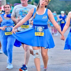 May include: Three people dressed as characters from the movie Toy Story are running in a race. The person in the center is wearing a blue dress with a yellow race bib that says "7113".