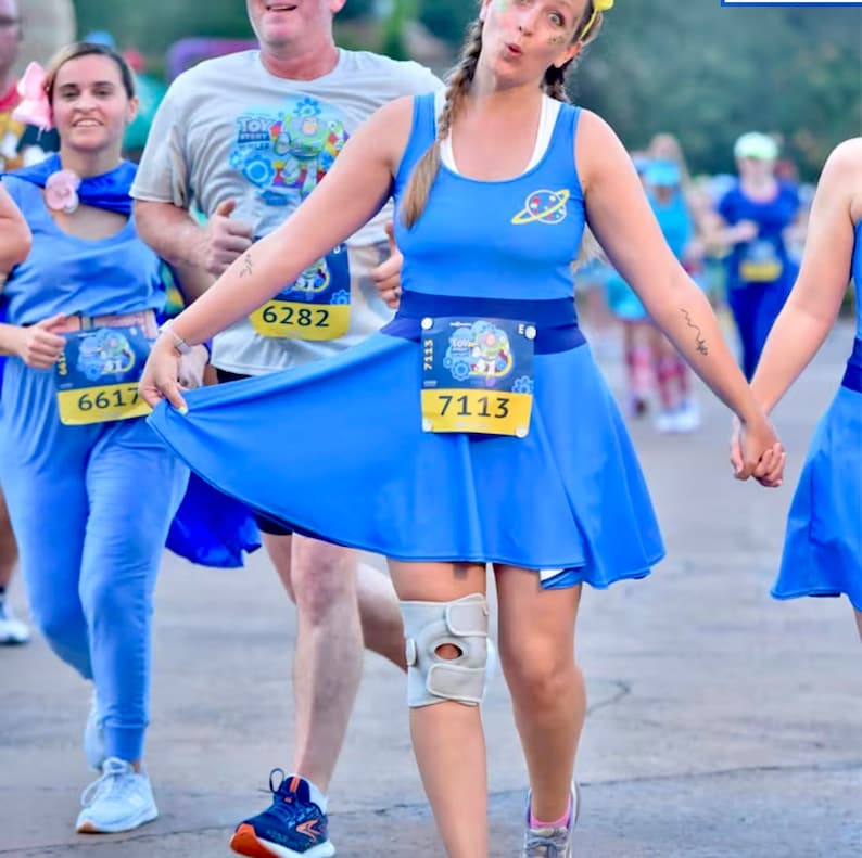 May include: Three people dressed as characters from the movie Toy Story are running in a race. The person in the center is wearing a blue dress with a yellow race bib that says "7113".