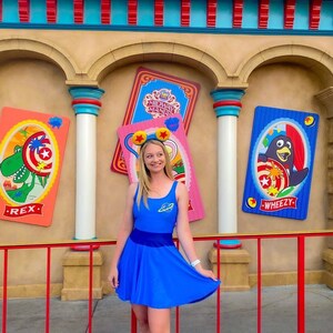 May include: A woman wearing a blue dress with a space-themed design stands in front of a colorful wall with Toy Story Midway Mania signs. The signs feature characters Rex and Wheezy.