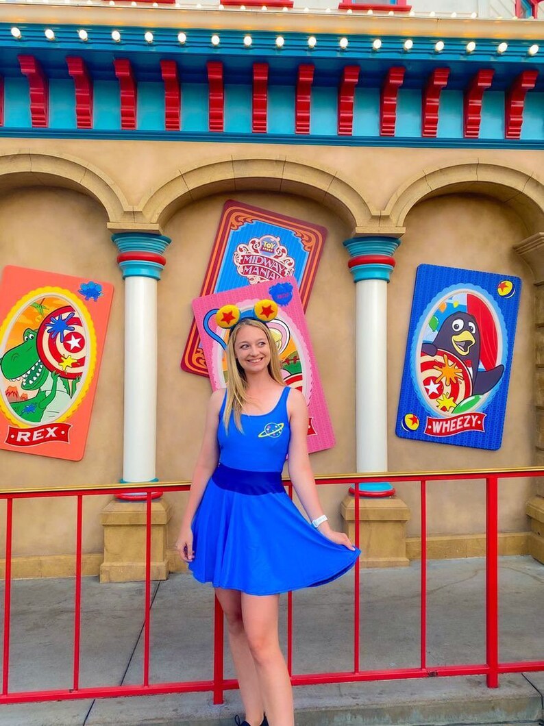 May include: A woman wearing a blue dress with a space-themed design stands in front of a colorful wall with Toy Story Midway Mania signs. The signs feature characters Rex and Wheezy.