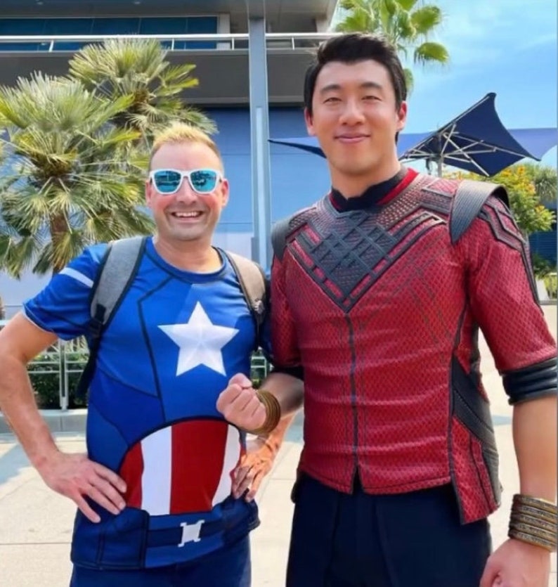 May include: Two people are standing next to each other. The person on the left is wearing a blue, white, and red t-shirt with a star and stripes design. The person on the right is wearing a red shirt with black accents and black pants.