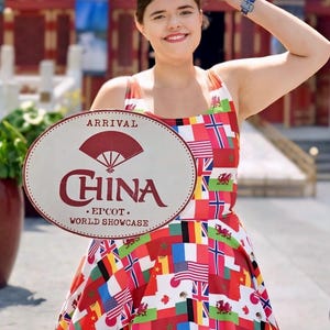 Puede incluir: Un vestido sin mangas con un estampado de varias banderas internacionales. El vestido se muestra junto a un letrero ovalado que dice ARRIVAL CHINA EPCOT WORLD SHOWCASE con un gr&aacute;fico de abanico rojo sobre la palabra CHINA.