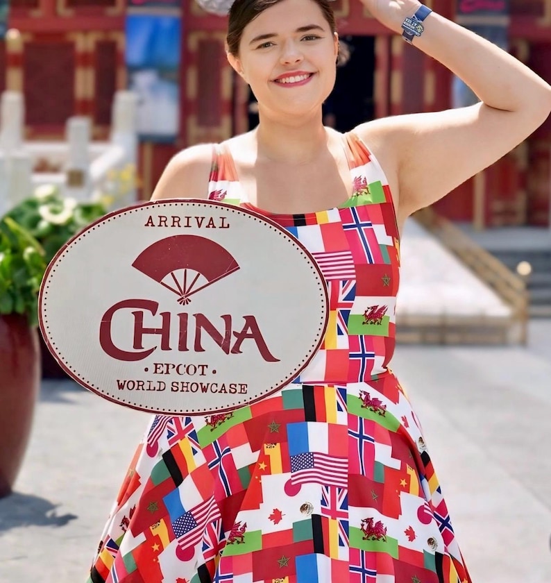 Puede incluir: Un vestido sin mangas con un estampado de varias banderas internacionales. El vestido se muestra junto a un letrero ovalado que dice ARRIVAL CHINA EPCOT WORLD SHOWCASE con un gr&aacute;fico de abanico rojo sobre la palabra CHINA.