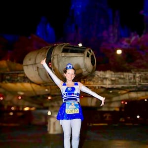 May include: A woman wearing a blue and white R2-D2 costume with a blue skirt and white leggings. She is holding a running bib with the number 22070. She is standing in front of a large spaceship.