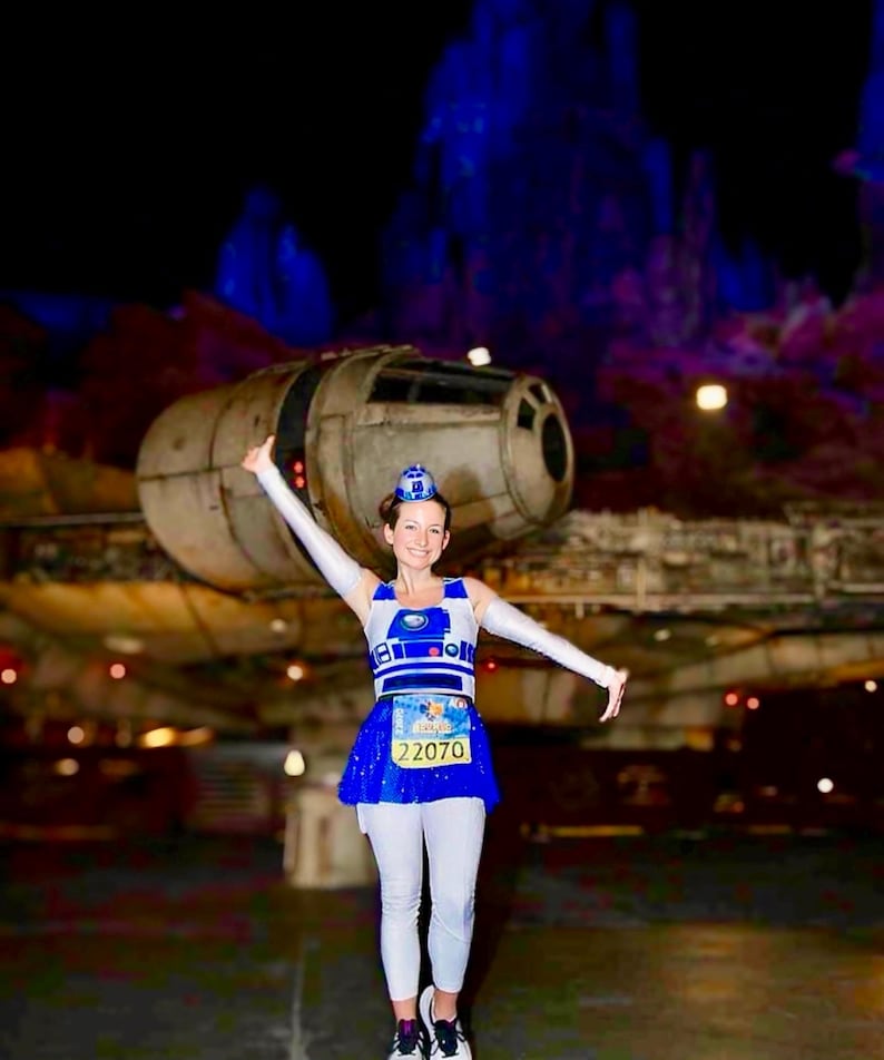 May include: A woman wearing a blue and white R2-D2 costume with a blue skirt and white leggings. She is holding a running bib with the number 22070. She is standing in front of a large spaceship.