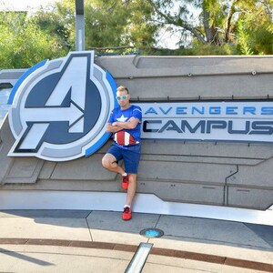 May include: A person wearing a blue shirt with a red, white, and blue design stands in front of a sign that says "Avengers Campus". The sign has a large, stylized "A" in the background.