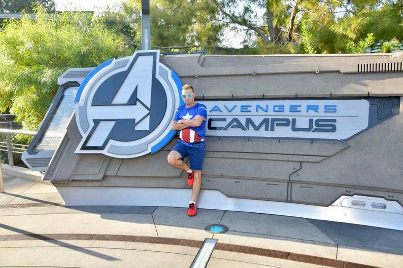 May include: A person wearing a blue shirt with a red, white, and blue design stands in front of a sign that says "Avengers Campus". The sign has a large, stylized "A" in the background.