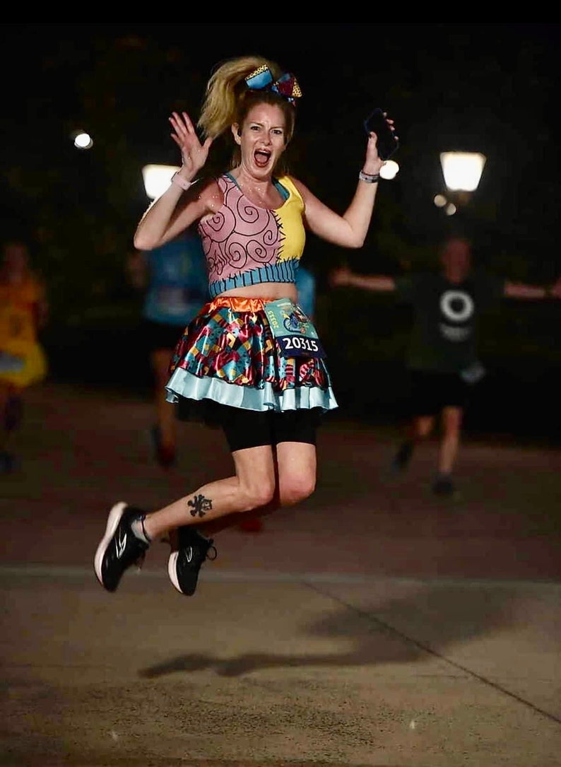 May include: A woman wearing a colorful, patterned skirt and a crop top with a cartoon design. She is wearing a race bib with the number 20315. She is jumping in the air with her arms raised and a big smile on her face.