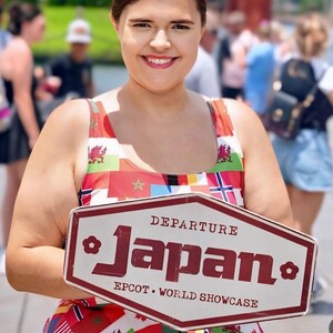Puede incluir: Un vestido sin mangas con un estampado multicolor de varias banderas internacionales. El vestido se combina con un letrero hexagonal blanco con borde granate que dice DEPARTURE Japan EPCOT WORLD SHOWCASE en letras de color granate.