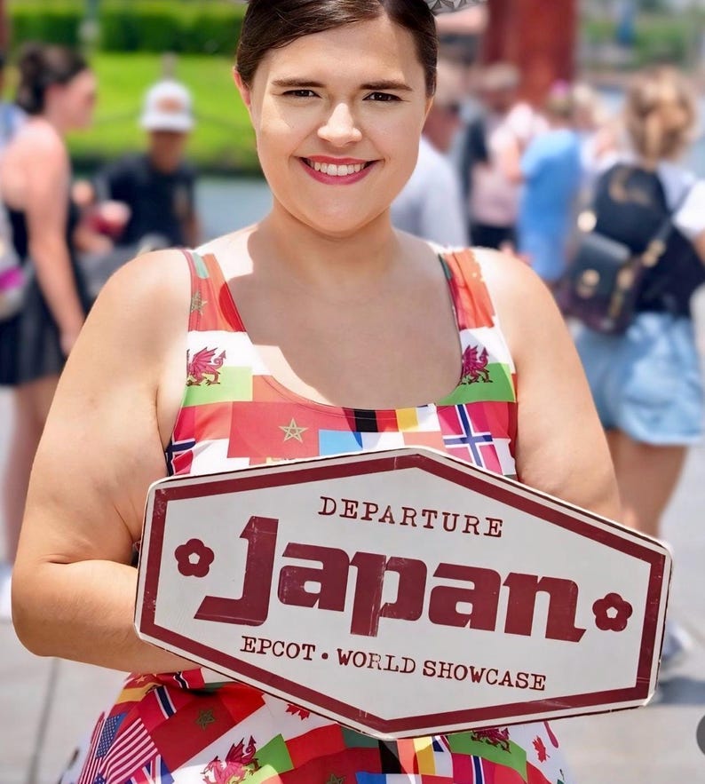 Puede incluir: Un vestido sin mangas con un estampado multicolor de varias banderas internacionales. El vestido se combina con un letrero hexagonal blanco con borde granate que dice DEPARTURE Japan EPCOT WORLD SHOWCASE en letras de color granate.