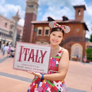 Puede incluir: Un vestido sin mangas con un patr&oacute;n colorido de varias banderas internacionales, combinado con una diadema de orejas de rat&oacute;n con un lazo central. El letrero que se sostiene muestra el texto ITALY DEPARTURE EPCOT WORLD SHOWCASE en letras rojas.