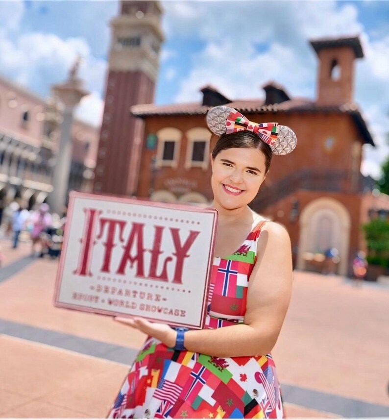 Puede incluir: Un vestido sin mangas con un patr&oacute;n colorido de varias banderas internacionales, combinado con una diadema de orejas de rat&oacute;n con un lazo central. El letrero que se sostiene muestra el texto ITALY DEPARTURE EPCOT WORLD SHOWCASE en letras rojas.