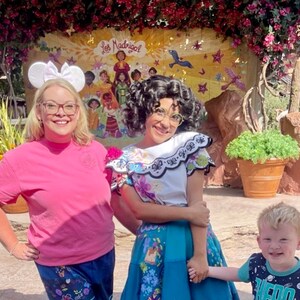 May include: A woman wearing a pink shirt and a Mickey Mouse headband poses with a woman dressed in a colorful traditional Mexican dress. The woman in the Mexican dress is holding hands with a young boy wearing a blue and black shirt with a space theme. The background features a mural with the text "Los Madrigal" and a colorful scene.