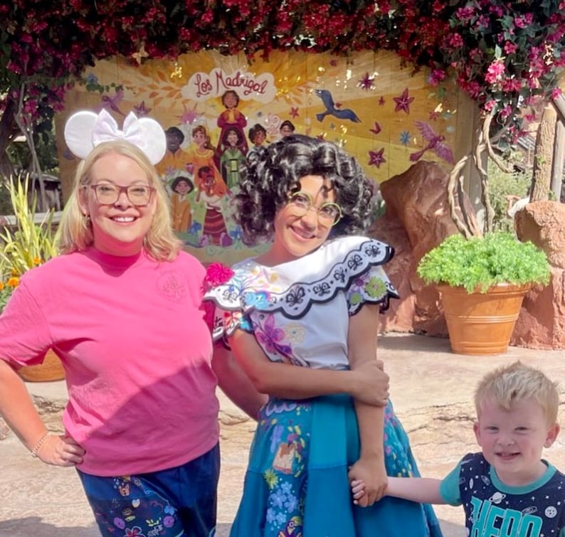 May include: A woman wearing a pink shirt and a Mickey Mouse headband poses with a woman dressed in a colorful traditional Mexican dress. The woman in the Mexican dress is holding hands with a young boy wearing a blue and black shirt with a space theme. The background features a mural with the text "Los Madrigal" and a colorful scene.