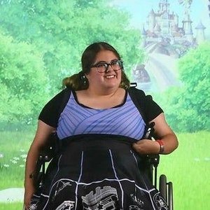 May include: A woman in a wheelchair wearing a blue and white striped top and a black skirt with a white graphic print. She is smiling and looking at the camera.