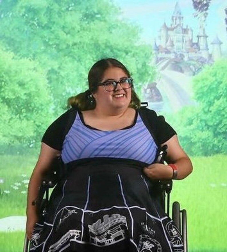 May include: A woman in a wheelchair wearing a blue and white striped top and a black skirt with a white graphic print. She is smiling and looking at the camera.