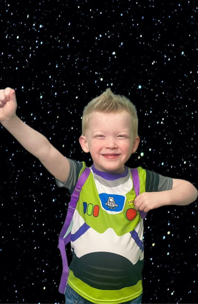 May include: A young boy wearing a green and white Buzz Lightyear costume with a purple backpack. He is smiling and has his arm raised in the air. The background is a black starry sky.