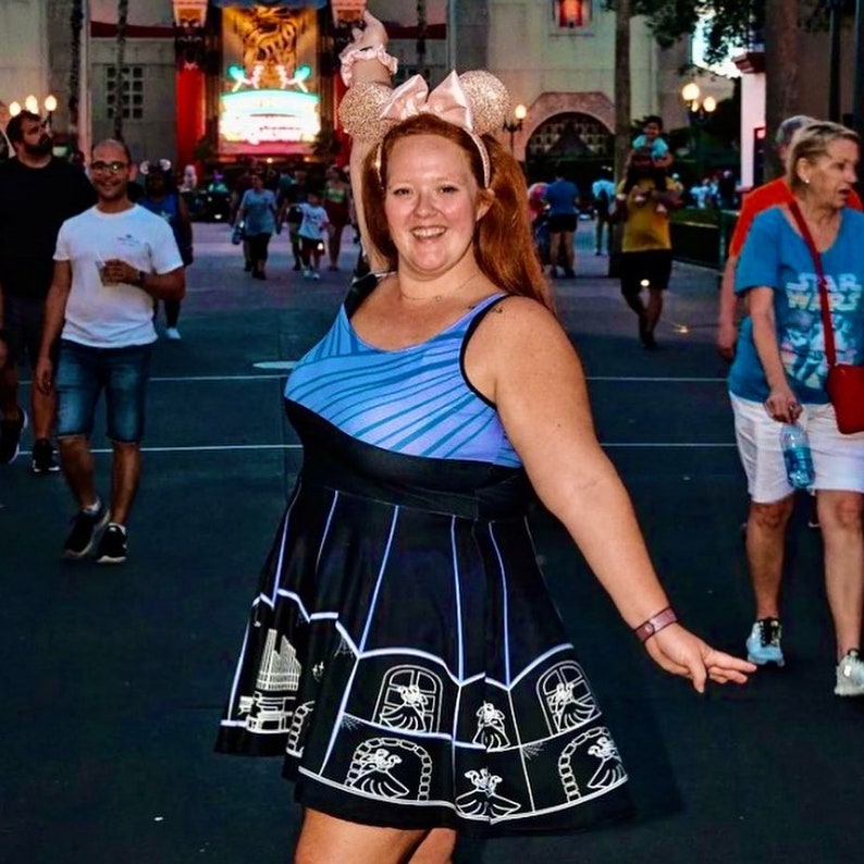 May include: A woman wearing a blue and black dress with a white graphic print of a building. She is wearing a pink headband with mouse ears and is smiling.