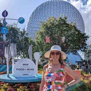 Puede incluir: Un vestido sin mangas con un patr&oacute;n colorido de varias banderas internacionales, usado con un sombrero de ala ancha de color claro y gafas de sol con montura roja, frente a una gran estructura esf&eacute;rica blanca y un letrero del Epcot Food & Wine Festival.