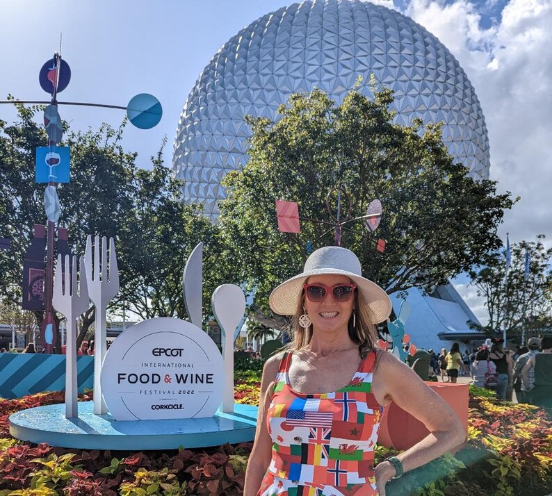 Puede incluir: Un vestido sin mangas con un patr&oacute;n colorido de varias banderas internacionales, usado con un sombrero de ala ancha de color claro y gafas de sol con montura roja, frente a una gran estructura esf&eacute;rica blanca y un letrero del Epcot Food & Wine Festival.