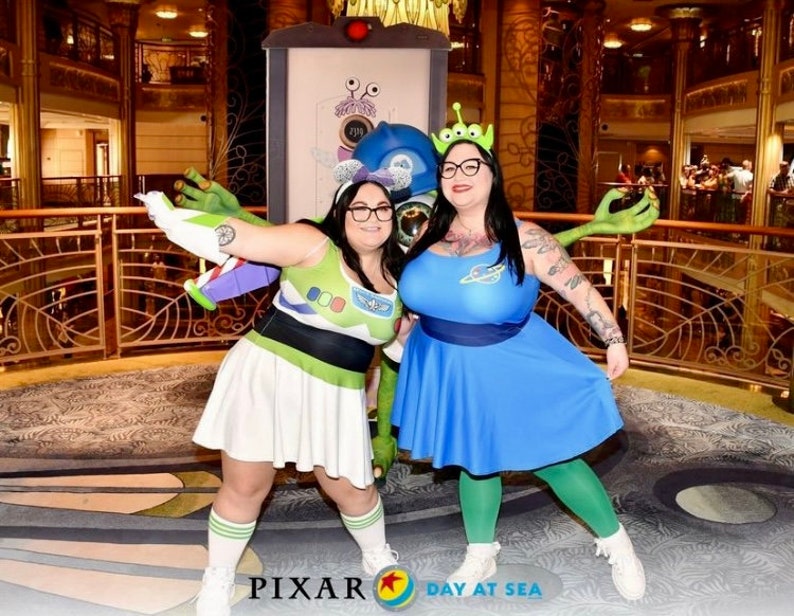 May include: Two people dressed as characters from the movie Toy Story. One person is dressed as Buzz Lightyear and the other is dressed as an alien. They are standing in front of a wall with a Pixar Day at Sea logo.