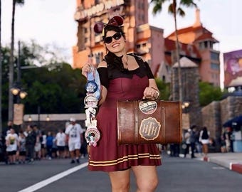 Puede incluir: Una maleta marrón de estilo vintage con el logotipo del Hollywood Tower Hotel y una pegatina circular, sostenida junto a una pila vertical de cuatro pines redondos de colección. La maleta tiene un acabado marrón oscuro con herrajes.