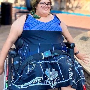 May include: A person in a wheelchair wearing a blue and black dress with a white graphic print. The dress has a black belt with a buckle. The person is smiling and looking at the camera.