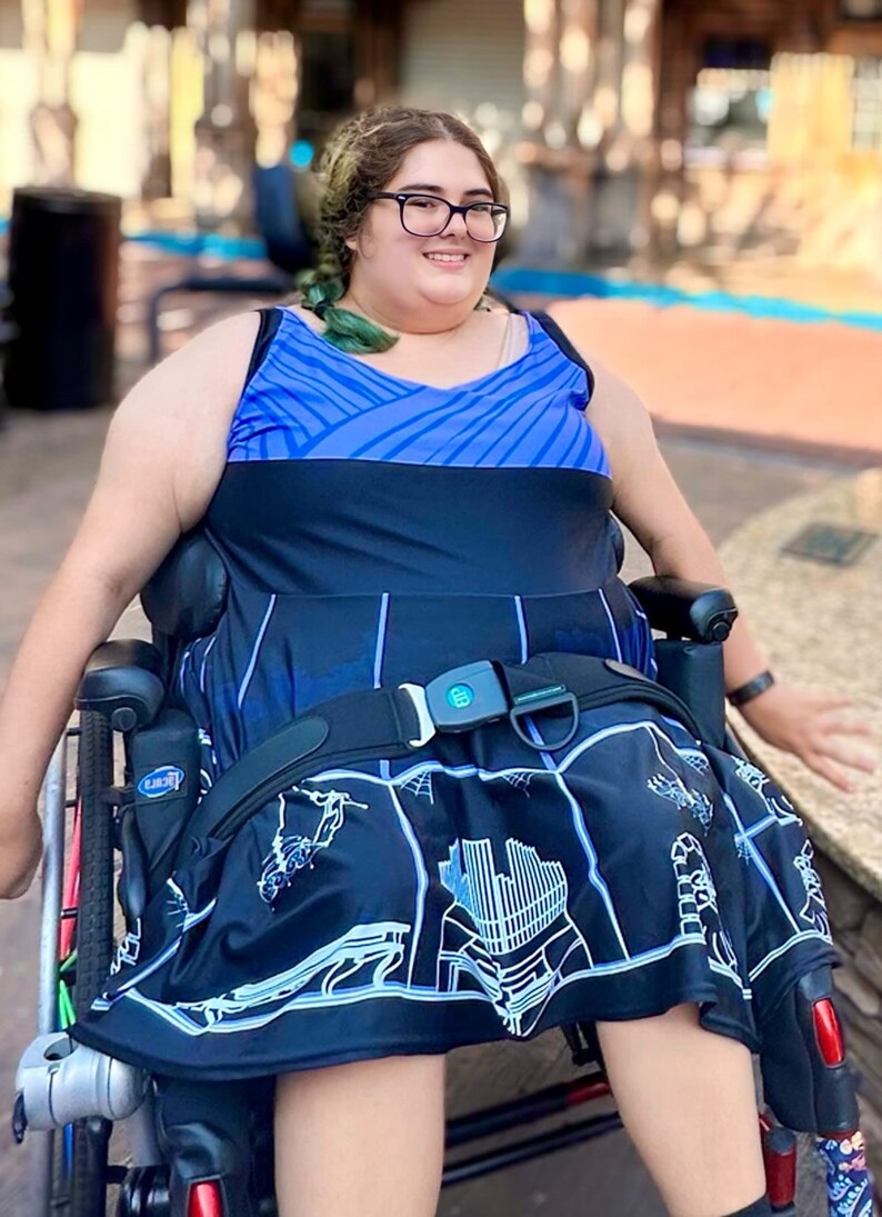 May include: A person in a wheelchair wearing a blue and black dress with a white graphic print. The dress has a black belt with a buckle. The person is smiling and looking at the camera.