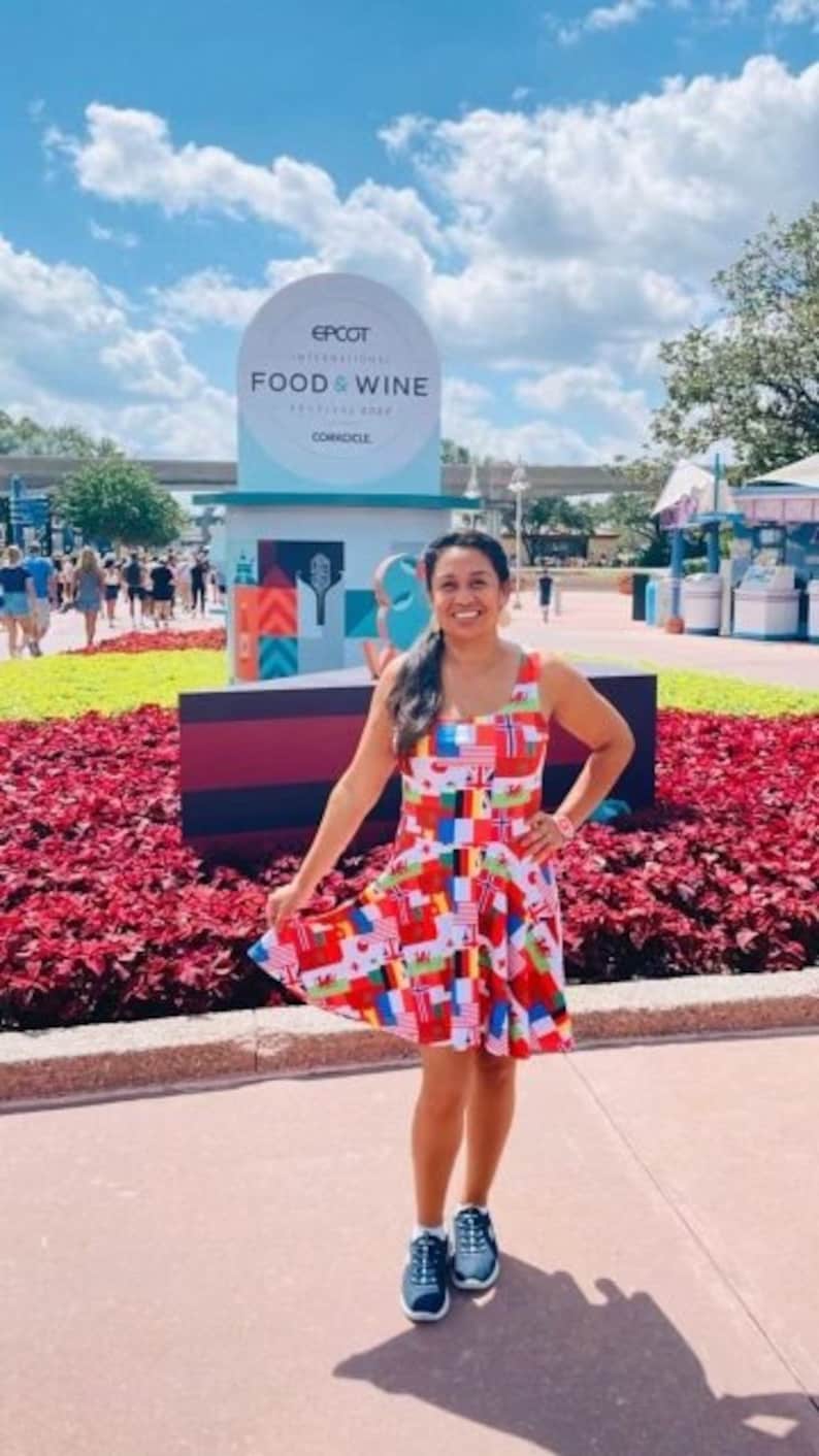 Puede incluir: Un vestido sin mangas hasta la rodilla con un estampado integral de varias banderas internacionales en colores vibrantes. El vestido tiene un escote redondo y una falda acampanada, combinado con zapatillas deportivas azul oscuro con suela blanca.