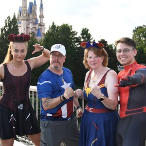 May include: Four people dressed as Marvel characters at a theme park. A woman in a red and black costume, a man in a blue and red Captain America costume, a woman in a blue and red Captain Marvel costume, and a man in a red and black Spider-Man costume.