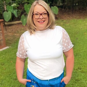 May include: A woman wearing a white t-shirt with polka dot mesh sleeves and blue shorts with a colorful floral print.