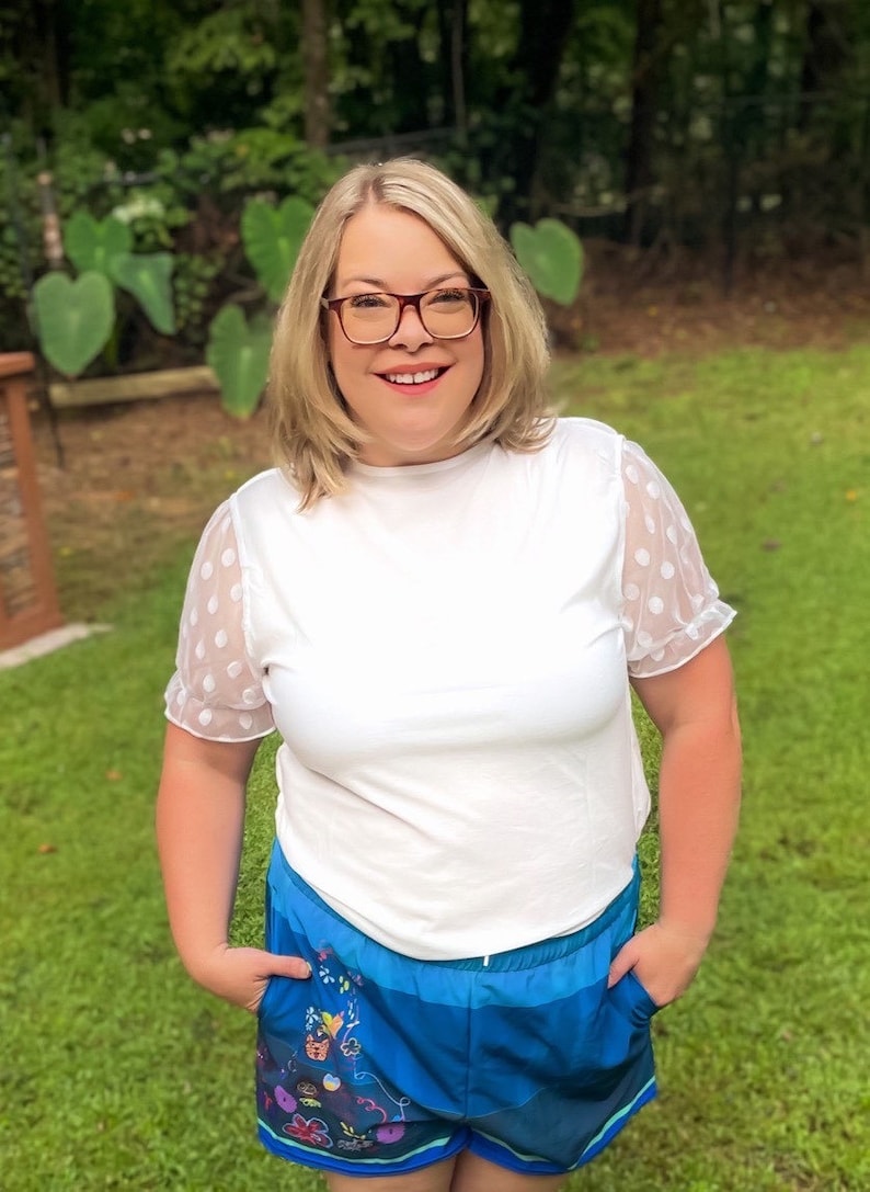 May include: A woman wearing a white t-shirt with polka dot mesh sleeves and blue shorts with a colorful floral print.