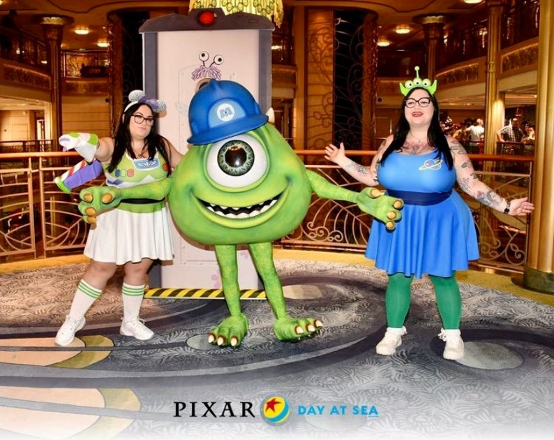 May include: Two people dressed as characters from the Pixar movie Monsters Inc. and Toy Story. One person is dressed as Mike Wazowski, the green one-eyed monster, and the other is dressed as a character from Toy Story. The background is a cruise ship with gold accents. The text "PIXAR DAY AT SEA" is visible at the bottom of the image.