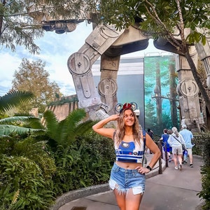 May include: A woman wearing a blue and white R2-D2 crop top and denim shorts stands in front of a large, grey, metal structure. She is wearing mouse ears with a red bow.