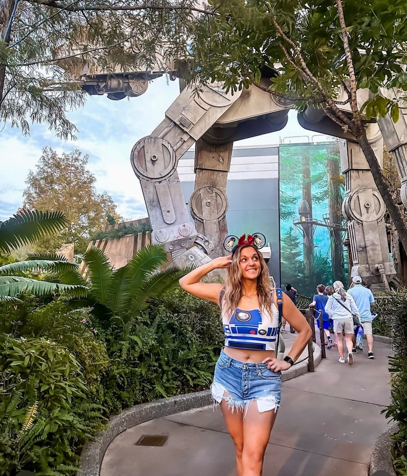 May include: A woman wearing a blue and white R2-D2 crop top and denim shorts stands in front of a large, grey, metal structure. She is wearing mouse ears with a red bow.