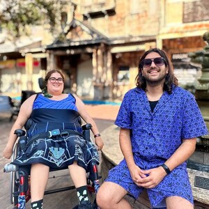 May include: Two people are sitting outside. One person is in a wheelchair wearing a blue and black patterned skirt and a blue top. The other person is wearing a purple shirt with a white pattern and black shorts. They are both smiling.