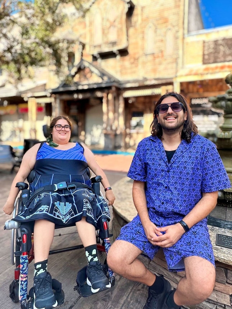 May include: Two people are sitting outside. One person is in a wheelchair wearing a blue and black patterned skirt and a blue top. The other person is wearing a purple shirt with a white pattern and black shorts. They are both smiling.