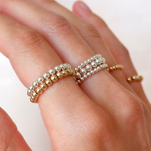 May include: A hand wearing four gold and silver beaded rings. The rings are stacked on two fingers.