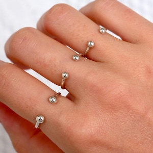May include: A set of four silver-toned open rings, each featuring a small, polished spherical bead at both ends. The rings are worn across four fingers, showcasing a simple, curved metal band design with a reflective, smooth metallic finish.