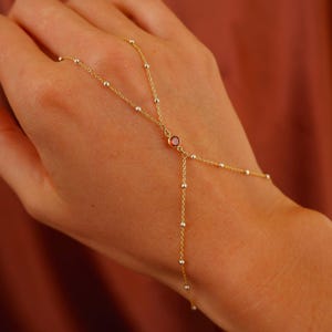 May include: A delicate gold hand chain with a small red gemstone at the center. The chain features small, evenly spaced beads and connects to a ring. The jewelry is displayed on a person's hand.