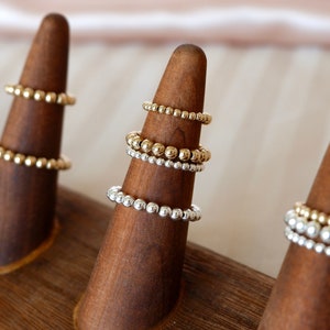 May include: A set of gold and silver beaded rings displayed on wooden ring holders. The rings are stacked on top of each other, creating a layered look.