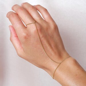 May include: A delicate gold hand chain. The jewelry piece features a ring that connects to a bracelet via a fine chain that drapes over the back of the hand. The hand chain is displayed against a soft, neutral background.